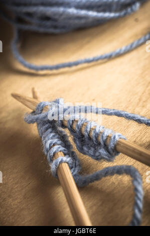 Ein gekreuztes paar Stricknadeln auf Holztisch mit Stichen im Gange mit blassen blauen Garn und Wolle Ball im Hintergrund. Leicht verblasst Stockfoto