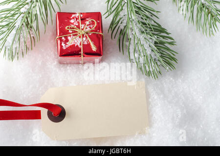 Overhead Schuss eine kleine Weihnachts-Geschenk-Box, mit leeren Paket Tag und Red Ribbon auf Kunstschnee und eine obere Grenze der Äste.  Das Label ist Stockfoto