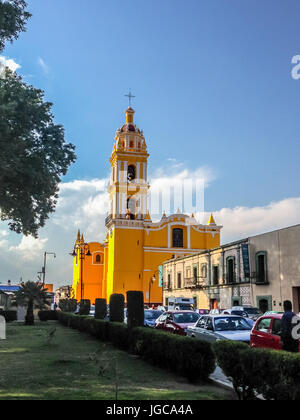 Leuchtend gelbe Fassade der Kirche San Apostolo auf dem Zocalo in Cholula, Mexiko betrachtet über Rasen und Sträuchern vor blauem Himmel Stockfoto