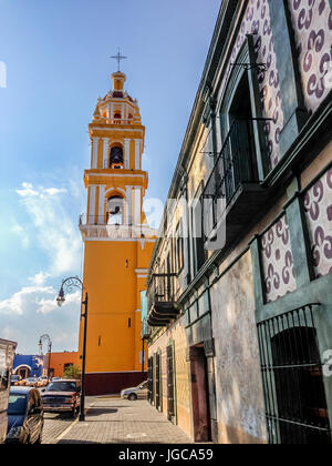 Leuchtend gelbe Fassade der Kirche San Apostolo auf dem Zocalo in Cholula, Mexiko betrachtet über Rasen und Sträuchern vor blauem Himmel Stockfoto