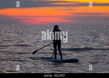 Silhouettierte Menschen stehen auf Paddelbrettern; Menschen paddeln beim Sonnenuntergang über der Irischen See, vom Badestrand des Resorts in Ainsdale, Merseyside, Großbritannien aus gesehen Stockfoto