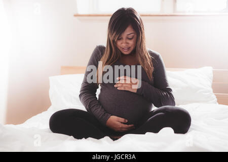 Schöne schwangere Frau auf Bett zu Hause sitzen. Junge schwangere Afrikanerin entspannend im Schlafzimmer mit ihren Händen auf den Bauch. Stockfoto