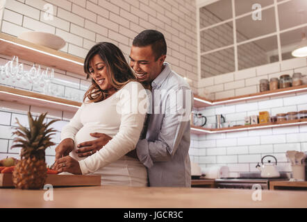 Junges Paar, gemeinsames Kochen in der Küche während Mann umarmt seine schwangere Frau. Glückliches junges Paar Kochen Essen zu Hause Hacken Gemüse auf kit Stockfoto