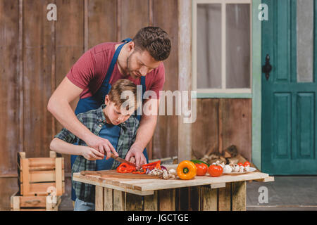 Vater in Schürze hilft Sohn Schneiden von frischem Gemüse im Freien, Vater und Sohn Kochen Konzept Stockfoto