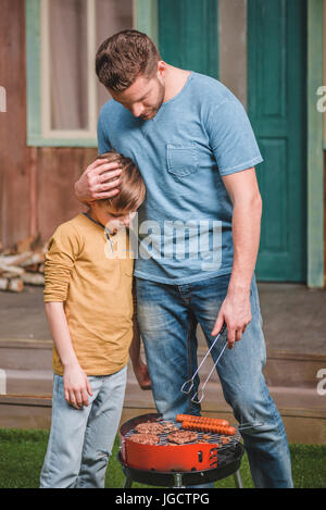 Vater und Sohn kochen Rindfleisch-Burger und Hot-Dog Würstchen auf dem Grill Stockfoto