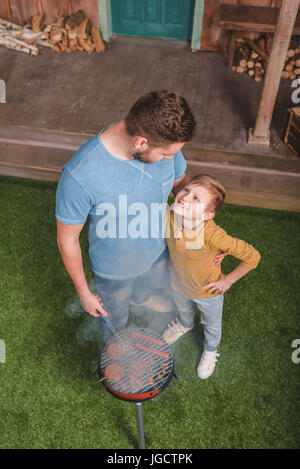 erhöhte Ansicht von Vater und Sohn kochen Rindfleisch-Burger und Hot-Dog Würstchen auf dem Grill Stockfoto