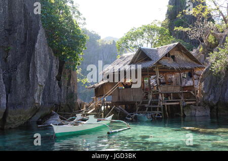 Kleine Holz-Ferienhaus / Hütte auf Stelzen und Felsen, in schönen blauen Lagune mit steilen Felswänden. Mensch in der Hängematte. Kleine Fischerboote. Coron Philippinen. Stockfoto