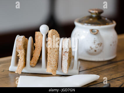 Vier Scheiben Toastbrot in Toast Halter auf Holztisch hautnah. Vordergrund Fokus, Teekanne in unscharfen Hintergrund. Niedrigen Winkel in der Nähe zu sehen. Shal Stockfoto