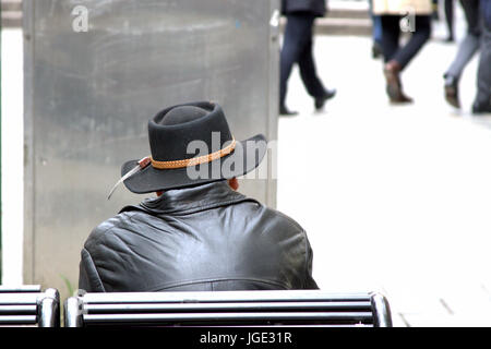 einziger Mann allein sitzen auf einer Bank in schwarz gekleidet, mit Leder Jacke und Cowboy-Hut Stetson Blick auf Menschen und die Welt geht durch Stockfoto