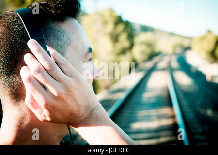 Nahaufnahme eines jungen Mannes gesehen von hinten mit Kopfhörern neben den Bahngleisen Stockfoto