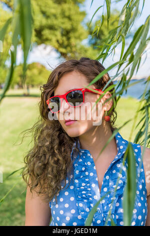 Porträt der jungen Frau in roten Sonnenbrillen stehen unter Weide im Sommer Stockfoto