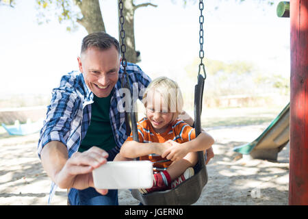 Vater nehmen Selfie mit Sohn sitzen auf Schaukel auf Spielplatz Stockfoto