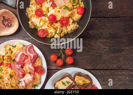 Rührei in einer Pfanne auf einen Teller und auf Brot, mit Cherry-Tomaten, Speck, Petersilie und Käse, geschossen von oben auf einem rustikalen Hintergrund Stockfoto