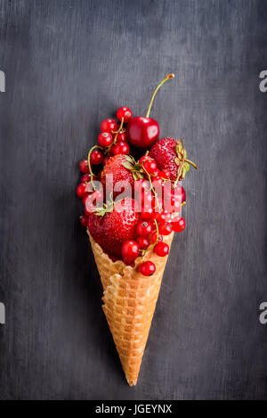 Frische rote Sommer Beeren - Erdbeeren, rote Johannisbeeren, Kirschen in das Waffelhörnchen in Form von Eis auf dem schwarzen Hintergrund aus Holz. Gesunden de Stockfoto