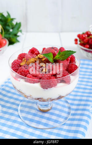 Cremige Süßspeise mit Frischkäse, Müsli, frische Himbeeren in eine Glasschüssel auf weißem Holz Hintergrund. Stockfoto