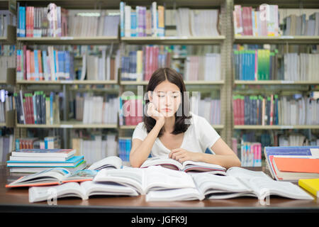 Asiatische Studentin langweilig Lesebuch in Bibliothek mit vielen Büchern in der Universität. Asiatische Studentin entmutigt Lesebuch für die Prüfung. Stockfoto