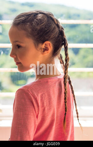 Mädchen mit niederländischen Zöpfen Blick aus Fenster im Profil. Kleines Kind mit braunen Haaren geflochten in einem rosa top Stockfoto