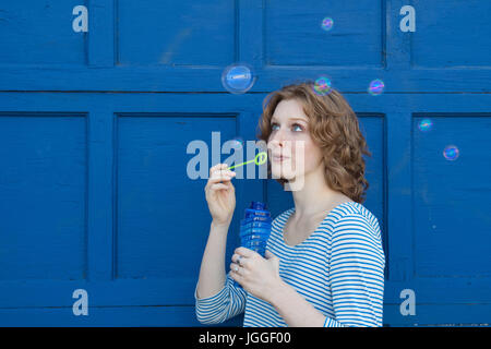 Rote Spitze Mädchen bläst Seifenblasen gegen blaue Wand Stockfoto