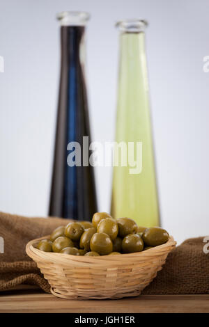 Nahaufnahme von grünen Oliven in Weidenkorb mit Ölflasche auf Tisch gegen Wand Stockfoto