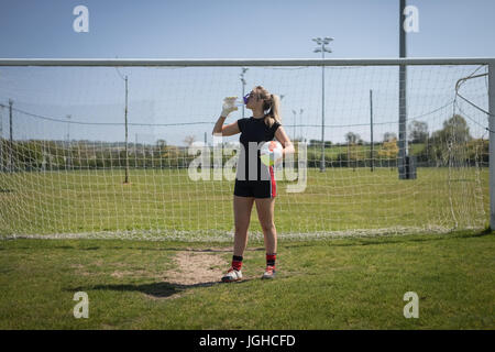 Gesamte Länge der weibliche Fußball Spieler Trinkwasser gegen Torpfosten auf Feld Stockfoto