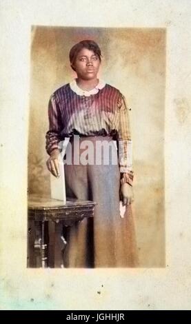 Drei Viertel Länge Portrait von afrikanische amerikanische Frau, mit einem leeren Gesichtsausdruck tragen ein dunkles Kleid, steht hinter dem Tisch, während sie 1920 eine weiße Buch auf dem Tisch steht. Hinweis: Bild wurde digital eingefärbt wurde mit einem modernen Verfahren. Farben können Zeit ungenau sein. Stockfoto