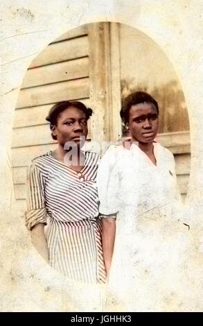 Drei Viertel Länge ovales Porträt von zwei jungen afro-amerikanischen Frauen, stehen vor einem Haus, das auf der linken Seite in einem gestreiften Kleid, die andere in einem weißen Kleid, sowohl mit angespannten Mimik, 1920. Hinweis: Bild wurde digital eingefärbt wurde mit einem modernen Verfahren. Farben können Zeit ungenau sein. Stockfoto