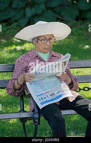 Ein asiatisch-amerikanische Frau in einem Schlapphut, lesen eine Zeitung im Washington Square Park in Manhattan, New York City. Stockfoto