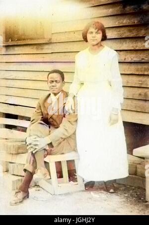 African-American Paar posieren vor einem Haus mit Holz Abstellgleis, Frau trägt weißes Kleid und stehend, Mann sitzt auf einer Holzkiste mit seiner Hand auf sein Knie, 1910. Hinweis: Bild wurde digital eingefärbt wurde mit einem modernen Verfahren. Farben können Zeit ungenau sein. Stockfoto