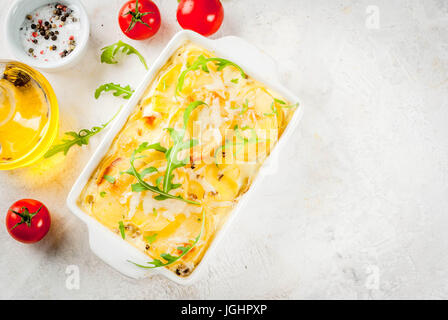 Kasserolle. Französische Küche. Hausgemachte Kartoffel-Gratin in einer keramischen Pfanne zum Backen. Auf weißem Marmor Stein Hintergrund. Mit Blättern frischer Rucola Stockfoto