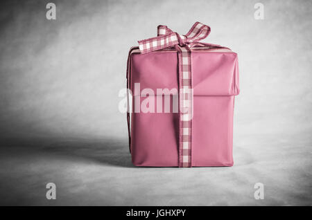 Eine einsame Geschenk-Box mit Deckel, in rosa Papier eingewickelt und mit einer Schleife Gingham (geprüft) Band gebunden.  Entsättigt grauen Pergament Hintergrund. Stockfoto