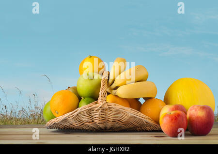 Eine Auswahl an Früchten, füllen einen Weidenkorb Blume und ein Überlaufen auf Holz beplankt Tabelle Wildgras Wiese mit blauen Sommerhimmel mit geringer Dichte Stockfoto