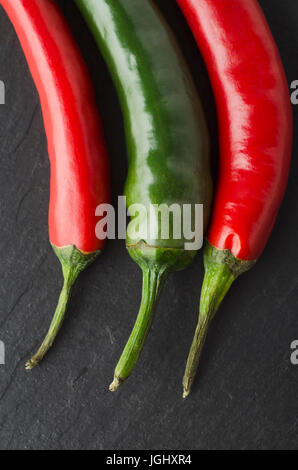 Nahaufnahme von drei Chilischoten auf schwarzem Schiefer von oben geschossen.  Grün in der Mitte, mit Paprika beiderseits. Stockfoto