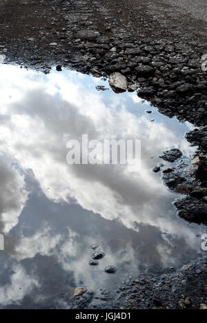 Risse und defekte Asphalt Schlagloch auf der Oberfläche einer bröckelnden Straße. Regen Wasser Pfütze sammelt sich in der gebrochenen Loch spiegelt den Himmel. Stockfoto