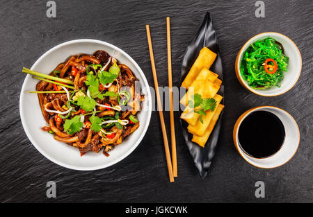 Asiatische Nudeln mit würziger Sojasauce, Hähnchenteile und Frühlingsrollen, Draufsicht, serviert auf schwarzem Stein. Stockfoto