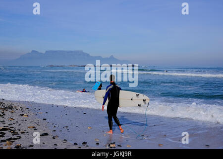 Kapstadt, Südafrika. Surfer genießen Sie einen frühen Winter morgen Surfen mit den Tafelberg in der Ferne in eine Schicht von Smog bedeckt. Stockfoto