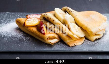 Leckere Crepes Dessert gefüllt mit Marmelade, Schokoladensauce, Banane, Pfirsich Scheiben und Puderzucker Stockfoto