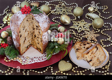 Schokolade Panettone Weihnachtskuchen mit Scheibe, Tand und Perle Golddekorationen, Stechpalme, Mistel, Efeu, Tanne und Kiefer Kegel auf Eiche Hintergrund. Stockfoto