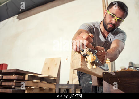 Eine junge Brünette mann Builder in Grün Brille und einem grauen T-Shirt Griffe eine Bar aus Holz mit einem Black Jack plane in der Werkstatt, im Hintergrund eine Menge Stockfoto