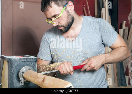 Eine junge Brünette Mann einen Builder in grün Schutzbrille, ein graues T-Shirt und einen Bart eine hölzerne Produkt auf einer Drehmaschine in der Werkstatt macht Stockfoto