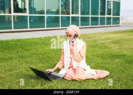 Eine junge ziemlich muslimische Geschäftsfrau in einem Park während des Gesprächs auf Smartphone Blick auf dem laptop Stockfoto