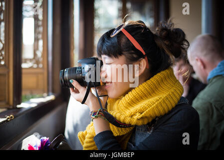 Junges Mädchen im gelben Schal, das mit einer DSLR-Kamera aus dem Fenster fotografiert. Stockfoto