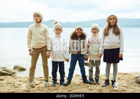 Gruppe von fröhlichen Kindern See hintereinander stehen, Zungen herausragen, während draußen spielen an warmen Herbsttag alle ähnlich gestrickte Kleidung gekleidet Stockfoto