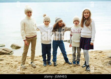 Gruppe von fröhlichen Kindern See hintereinander posiert spielerisch an Kamera auf warmen Herbsttag in ähnlich gestrickte Kleidung gekleidet stehen Stockfoto