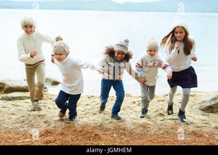 Gruppe von glücklichen Kindern spielen im freien laufen am Seeufer an warmen Herbsttag, die alle gekleidet ähnlicher Kleidung stricken Stockfoto