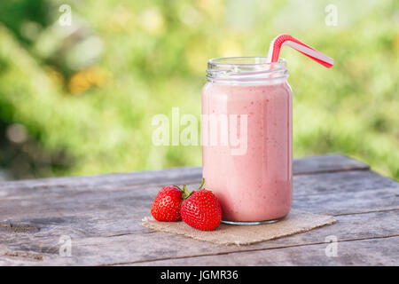 Erdbeer Milchshake in Glas und frischen Reifen Erdbeeren auf Holztisch mit grünen Natur der Hintergrund jedoch unscharf. Rosa-Smoothie. Gesunden Sommer Getränk con Stockfoto