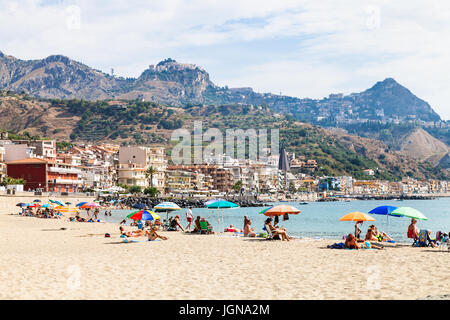 GIARDINI NAXOS, Italien - 28. Juni 2017: Menschen am Sandstrand in Giardini-Naxos-Stadt und Blick auf Taormina Stadt am Kap. Giardini Naxos ist Badeort Stockfoto