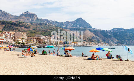 GIARDINI NAXOS, Italien - 28. Juni 2017: Touristen am Sandstrand in Giardini-Naxos-Stadt und Blick auf Taormina Stadt am Kap. Giardini Naxos ist am Meer reso Stockfoto