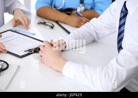 Zwei Ärzte und männlicher Patient diskutieren Krankengeschichte am Tisch, Nahaufnahme Stockfoto