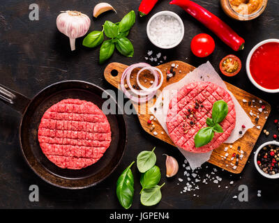 Rohes Rindfleisch Fleisch Steak Schnitzel für Burger in Pfanne, Schnitzel auf Holzbrett mit Gewürzen, Gemüse, Saucen, Ketchup und Senf auf schwarz Beton zurück Stockfoto