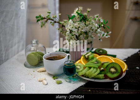 Frühstück mit Tee oder Kaffee mit Orange und Kiwies Scheiben und grüne Makronen auf einer weißen Decke mit Kirschblüten Stockfoto